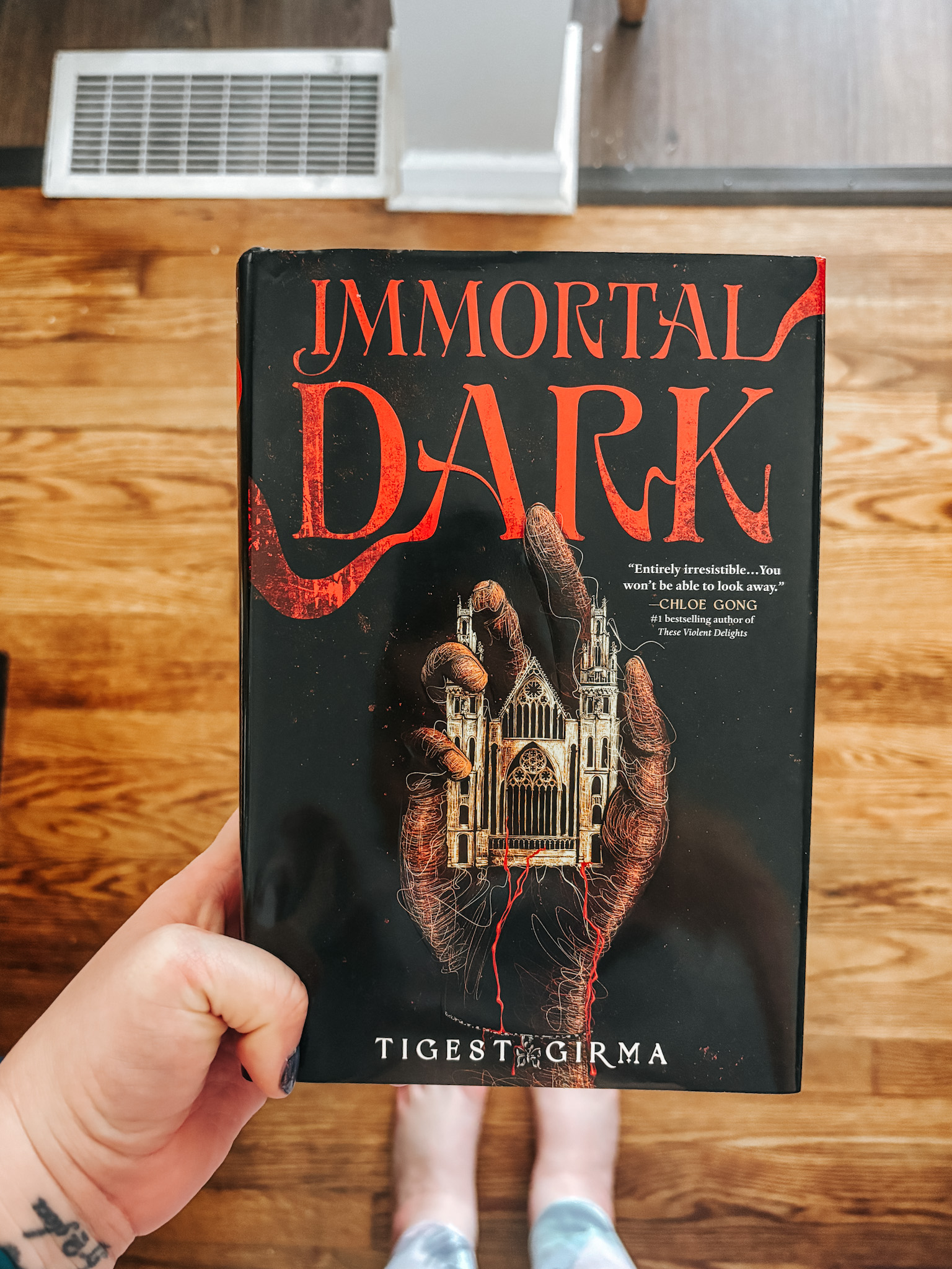 A white woman holds the book Immortal Dark over her feet while standing on a wooden floor.